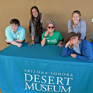 A group of Day Camp Teachers behind a table with a turquoise cloth imprinted with the Desert Museum logo in white