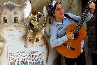 Educator Michelle plays guitar next to the entrance to Packrat Playhouse